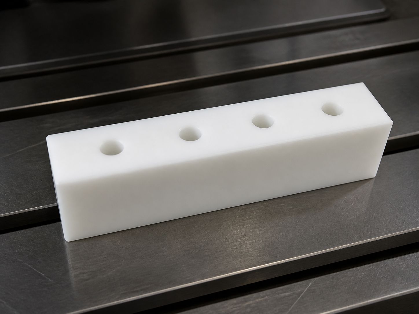 A thick white wear bar with four drilled holes resting across the T-slots of a machine table.