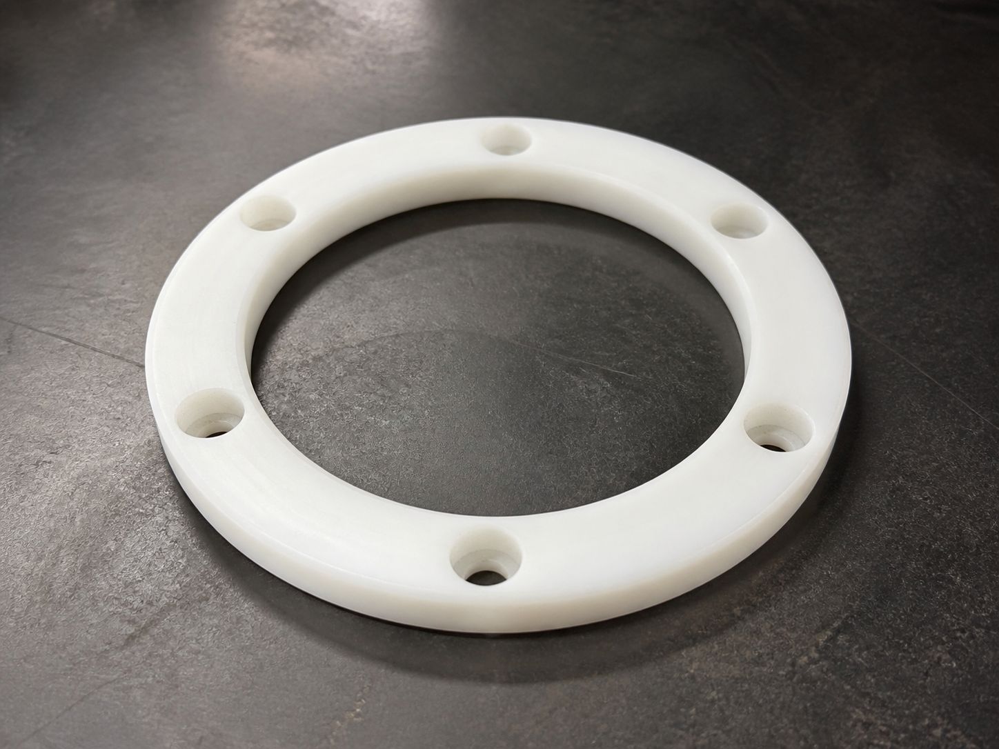 A white machined wear ring with six bolt holes, seen from above on a steel bench.